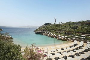Idyllischer Strandabschnitt am Maxx Royal Bodrum mit kristallklarem Wasser, eleganten Sonnenliegen, grüner Umgebung und Blick auf eine felsige Landzunge mit Skulptur – exklusiver Rückzugsort an der türkischen Riviera.