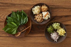 holzschüsseln mit frisch geschnittenen zutaten wie zeiwbeln ananas kraut nüsse chillis und weiteres gemüse daneben blätter und eine sauce zum kochen mit holzlöffel auf einem holz tisch im schönen exklusiven klassischen urigen und altmodischen hotel phun b