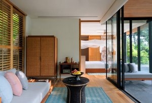 foto von einer pool villa suite blick aus dem wohnzimmer mit holz schrank in der ecke und sofa und kleinem kaffe tisch mit einer gefüllten obst schale auf dem tisch im hintergrund blick ins schlafzimmer mit edlen himmelbett und ventilator an der decke die