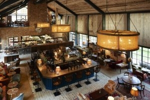 oben ansicht auf das klassische restaurant im country style mit einer grossen holz bar und barhockern rund herum daneben esstische und oben eine sitzgelegenheit mit weiteren tischen zum essen das restaurant hat eine ziegelstein wand und ein hohes spitzes 